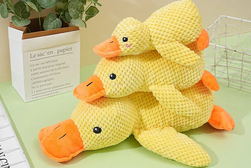Three yellow plush Dog Toy ducks sitting on a green surface with a plant and basket in the background.